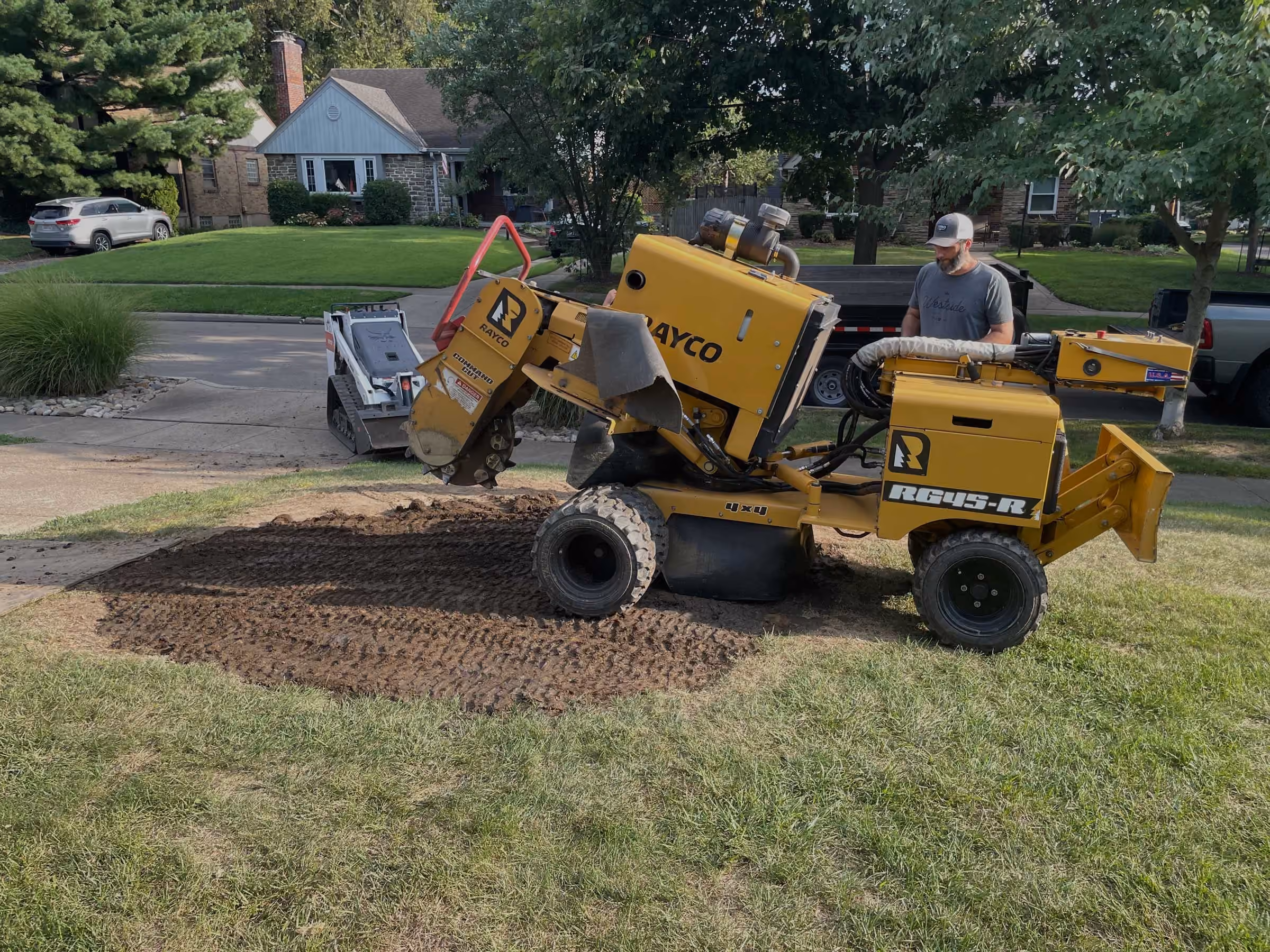 A Cincinnati Stump Grinding crew member beside a yellow Rayco stump grinder in a front yard, with the ground-out stump area in front of it